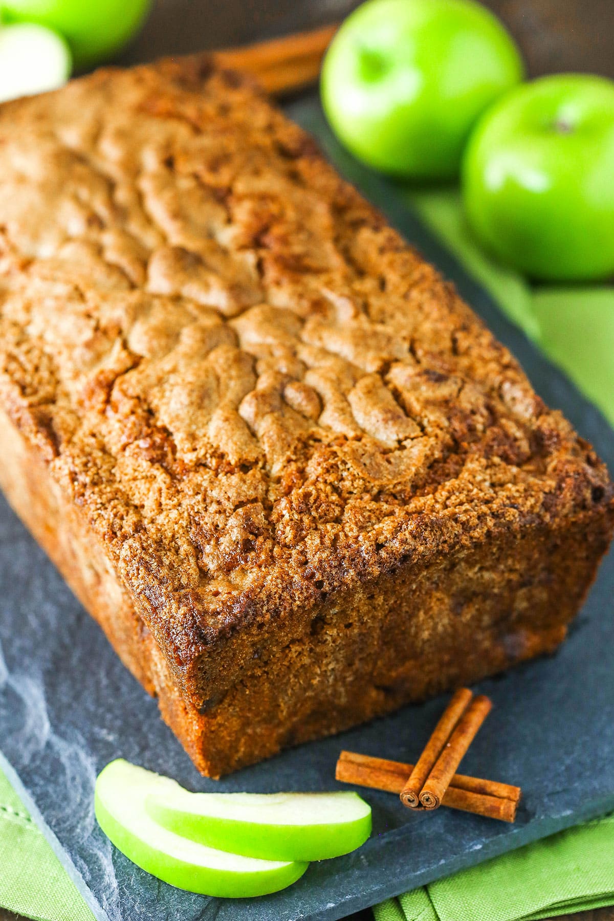A loaf of Apple Bread on a black cutting board with green apples in the background