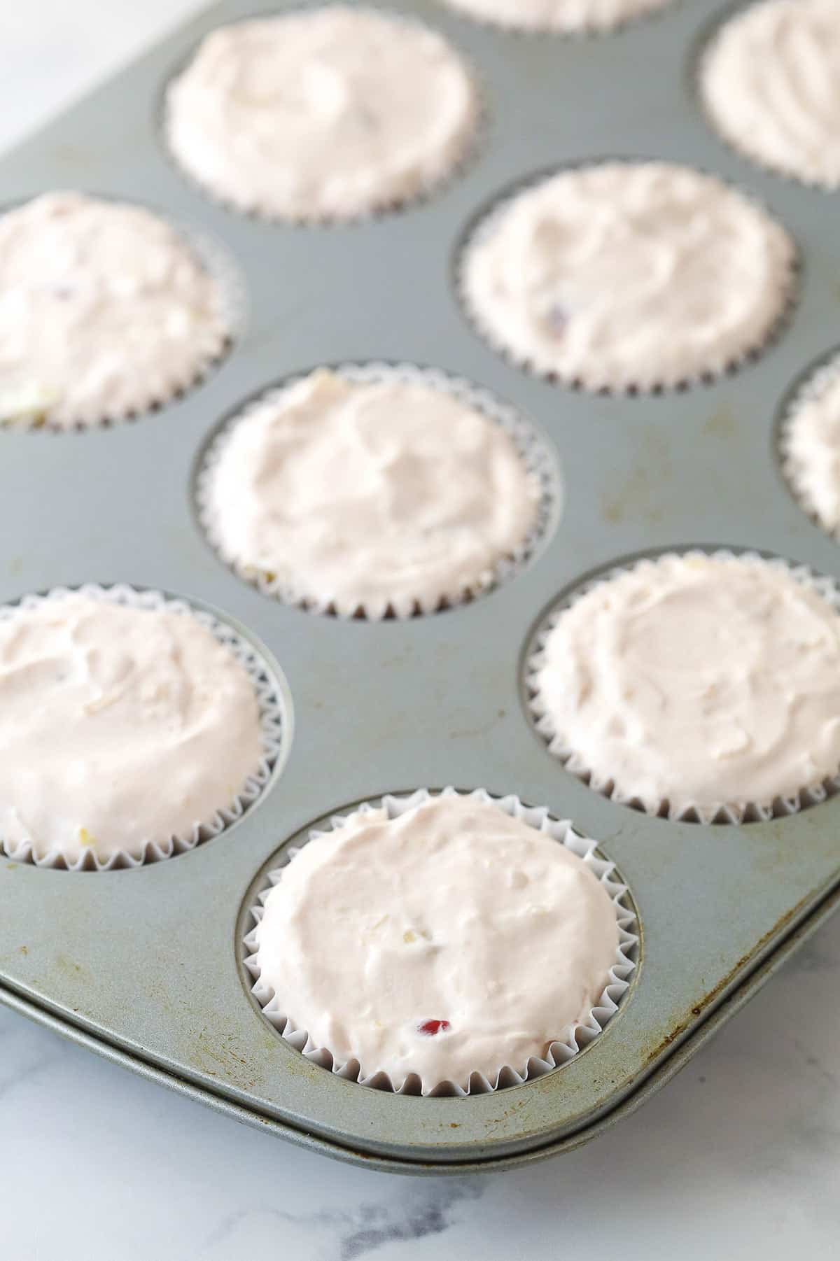 A cupcake pan lined with cupcake liners filled with creamy fruit salad ready to freeze.