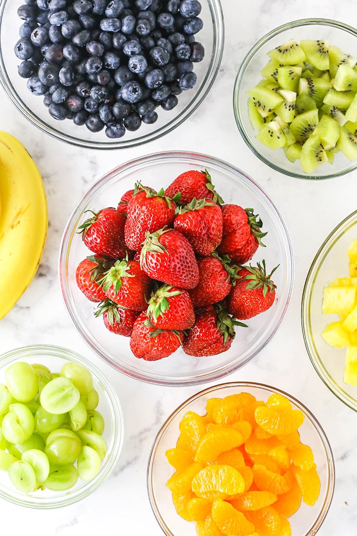 all the different chopped fruits in separate bowls - strawberries, blueberries, kiwi, grapes, oranges, pineapple and bananas