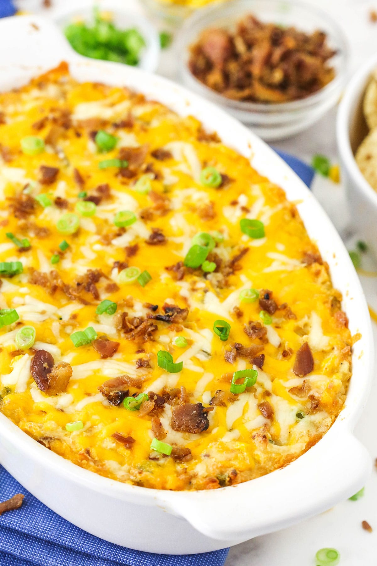 A serving dish full of cheesy Mexican-style dip beside a bowl of bacon bits and chopped green onions