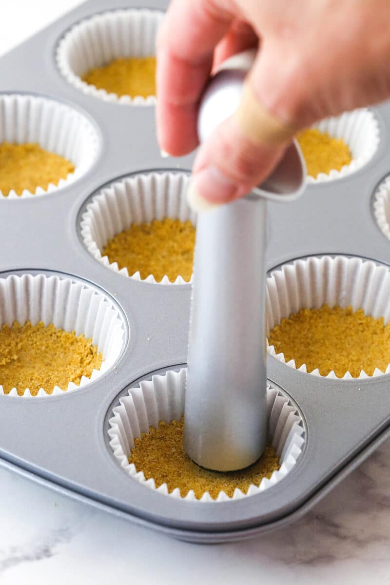 Pressing graham cracker crust into the bottoms of lined muffin tins.