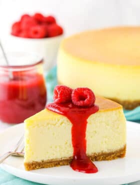 Slice of New York Style Cheesecake topped with raspberry sauce and raspberries on a white plate with a fork.
