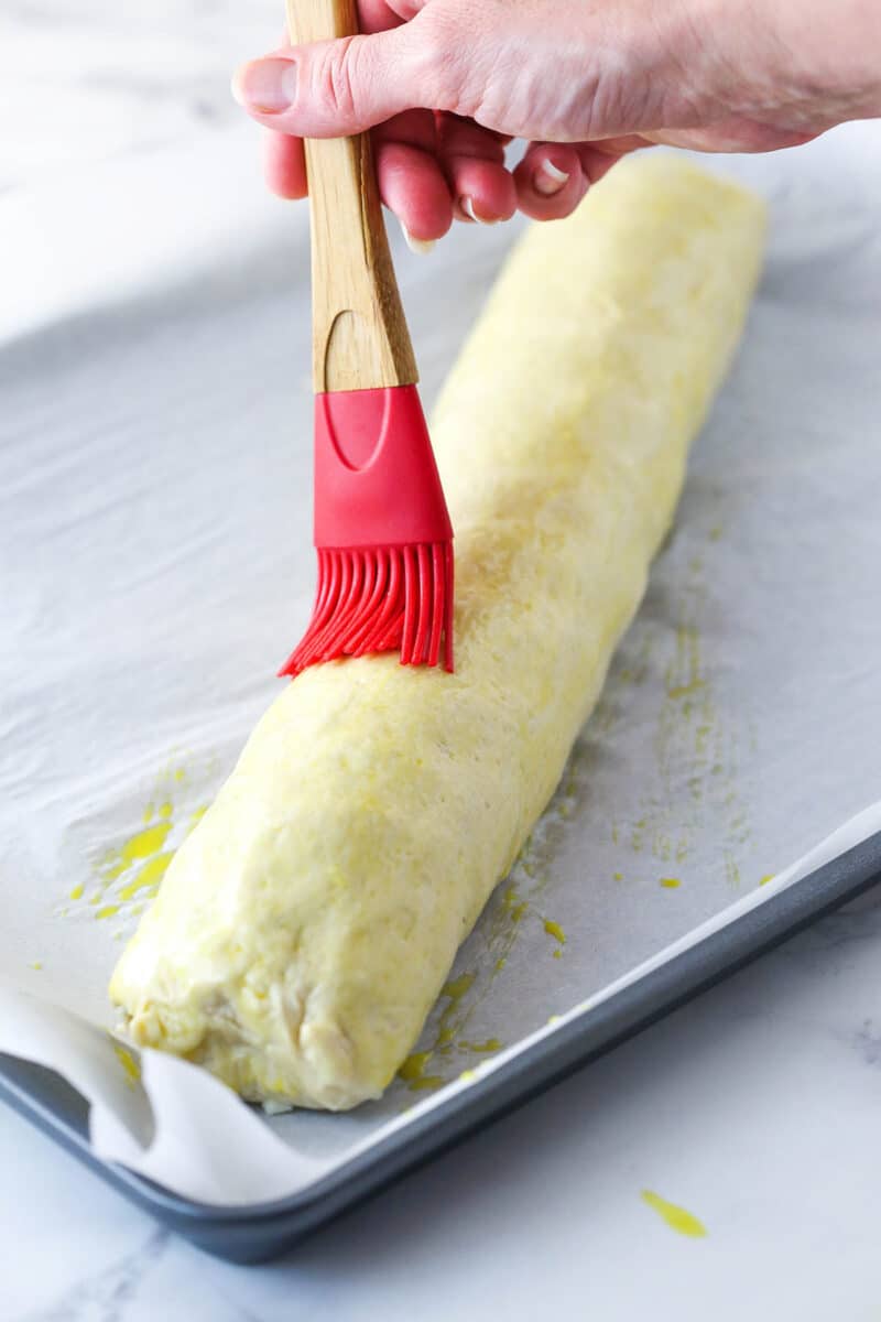 Brushing egg wash over an unbaked pepperoni roll on a baking sheet lined with parchment paper.