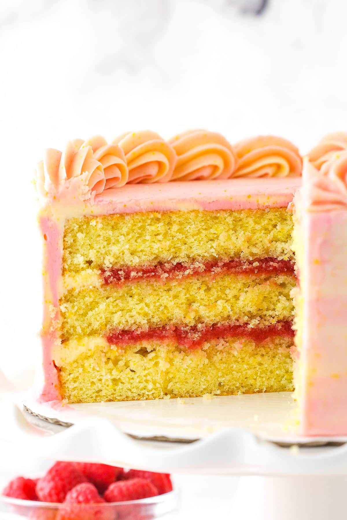 Raspberry orange layer cake on a cake stand with a slice cut out of it near a bowl of raspberries.