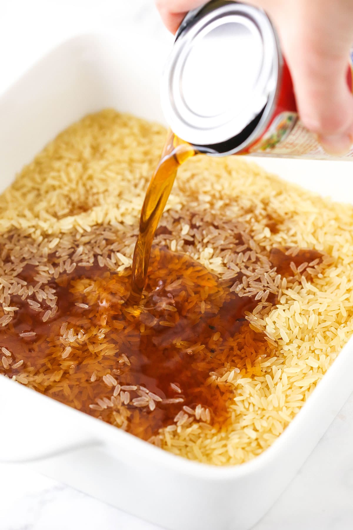 A can of condensed soup being poured into a baking dish full of long-grain rice