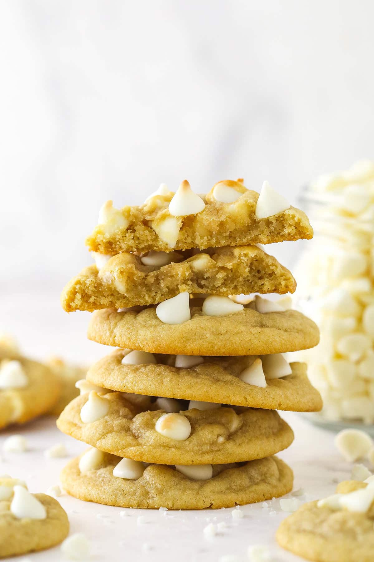A stack of white chocolate chip cookies surrounded by other white chocolate chip cookies and with the top cookie broken in half.